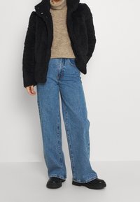Chaqueta de piel sintética negra, suéter de cuello alto beige, pantalones vaqueros de pierna ancha azules y botas negras gruesas. La textura suave y el ajuste relajado son características destacadas.