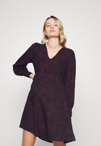 Vestuário preto com mangas longas, decote em V, apresentando padrões ondulados em roxo. A barra é ampla e possui um acabamento texturizado.