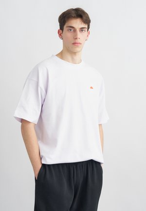 Giovane uomo con capelli castani corti che indossa una maglietta bianca oversize semplice e pantaloni neri, in piedi con le mani nelle tasche.