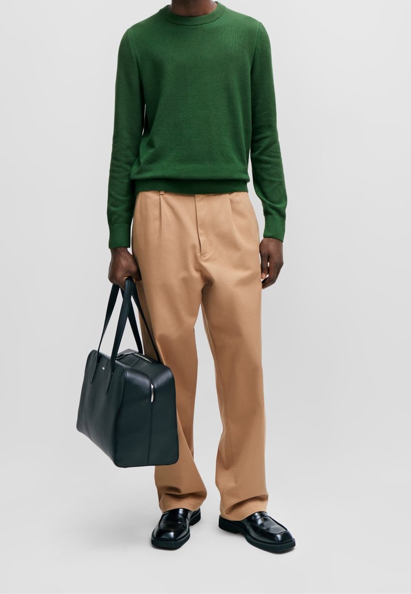 Pull à manches longues vert, pantalon plissé beige, chaussures en cuir noires et un sac à main vert foncé avec des poignées. Textures lisses, design simple.