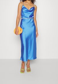 Blue satin slip dress with cowl neck, embellished waist, and spaghetti straps, paired with a circular orange clutch and green strappy heels.