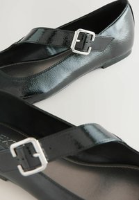 Black patent leather flats with a wide strap and a silver buckle. Features a smooth, shiny texture and a low-profile design.