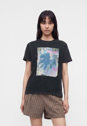 Femeie purtând un tricou negru cu o grafică cu palmier și text, asortat cu pantaloni scurți maro în carouri, stând în fața unui fundal alb simplu.