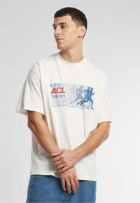 White cotton t-shirt with short sleeves, featuring a graphic print in blue and red that includes running figures and text.