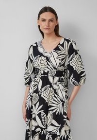 Zwarte jurk met wit loofpatroon, V-hals, elastische taille en korte pofmouwen. Gelaagde rok met contrasterende texturen en accenten.