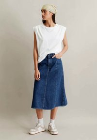 Frau mit beigem Kopftuch, ärmellosem weißen Oberteil, blauer Denim-Midiskirt, weißen Socken und beige-weißen Sneakers, steht vor einem einfarbigen Hintergrund.