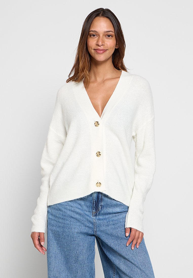 Cardigan bianco a coste con scollo a V, maniche lunghe e tre bottoni dorati sul davanti. Texture morbida, vestibilità leggermente oversize.
