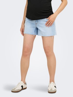 Vrouw draagt lichtblauwe denim shorts, zwart topje en witte sneakers met zwarte strepen, staande met één hand in haar zak.