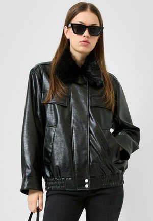 Junge Frau trägt schwarze Kunstlederjacke mit Fellkragen und schwarze Sonnenbrille, hält Handtasche, vor schlichtem hellem Hintergrund.