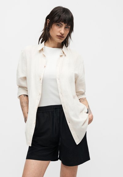 Γυναίκα με σκούρα μαλλιά φοράει ένα λευκό πουκάμισο κουμπωτό πάνω από ένα λευκό τοπ και μαύρα σορτς, στέκεται με τα χέρια στις τσέπες με φόντο ένα μονόχρωμο υπόβαθρο.
