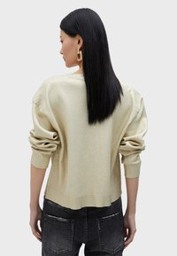 Pullover in oro chiaro con maniche lunghe, vestibilità rilassata, tessuto testurizzato e polsini a coste, indossato sopra jeans scuri grigi con effetto usurato.