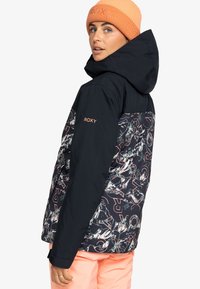 Schwarze und gemusterte Jacke von Roxy mit Kapuze, die ein farbenfrohes abstraktes Design aufweist, kontrastiert mit einer korallefarbenen Mütze.