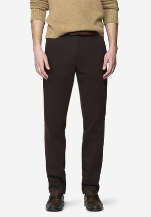Man draagt donkerbruine broek, bruine leren riem, beige gebreide trui met opgerolde mouwen en bruine leren loafers staand.
