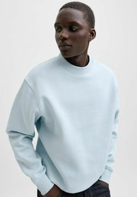 Hellblauer Pullover mit hohem Kragen, lockerer Passform und langen Ärmeln. Der Stoff wirkt weich mit einer glatten Textur. Auffällige Nahtdetails sind sichtbar.