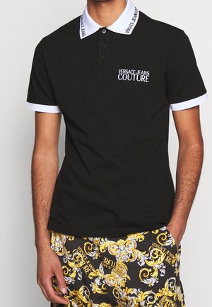 Homme portant un polo noir avec col et poignets blancs, logo "Versace Jeans Couture", et pantalon noir, or et blanc à motifs avec la même marque.