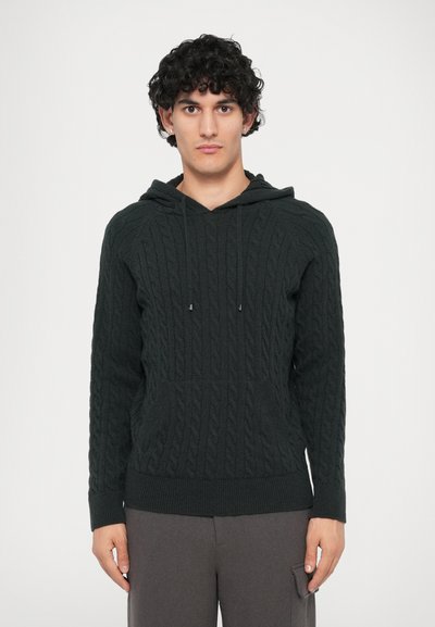 Felpa con cappuccio a coste di colore verde scuro, dotata di tasca frontale, cappuccio con cordoncino, orlo a coste e motivo testurizzato. Abbinata a pantaloni grigi.