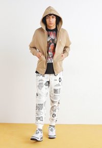 Sudadera beige con cremallera y capucha, camiseta gráfica negra, pantalones de chándal blancos con estampado y zapatillas grises con acentos negros.