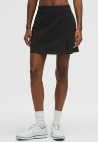 Falda pantalón negra con textura suave, que presenta una abertura frontal y una cintura plana. Combinada con zapatos de golf blancos y calcetines de longitud media.