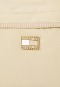 Borsa in tessuto beige con una patch rettangolare in tono oro che riporta la scritta "TOMMY JEANS", con una texture liscia e cuciture a contrasto.