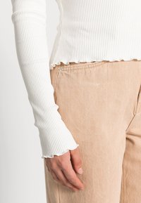 Weißes, geripptes Langarmoberteil mit rüschiertem Saum, kombiniert mit hellbeigen Hosen, die sichtbare Taschensteppnähte und eine weiche Textur aufweisen.
