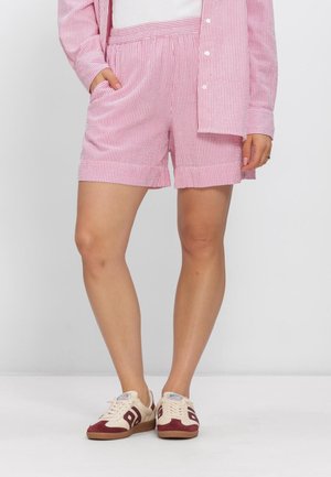 Person, die rosa-weiß gestreifte Shorts und ein passendes offenes Hemd mit weißen Turnschuhen trägt, steht vor einem einfarbigen Hintergrund.