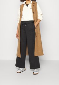 Lång brun ullväst över en vit dragkedja-fleece och svarta dragkedjebyxor. Fötter i silverfärgade sandaler. Avslappnad, vid passform.