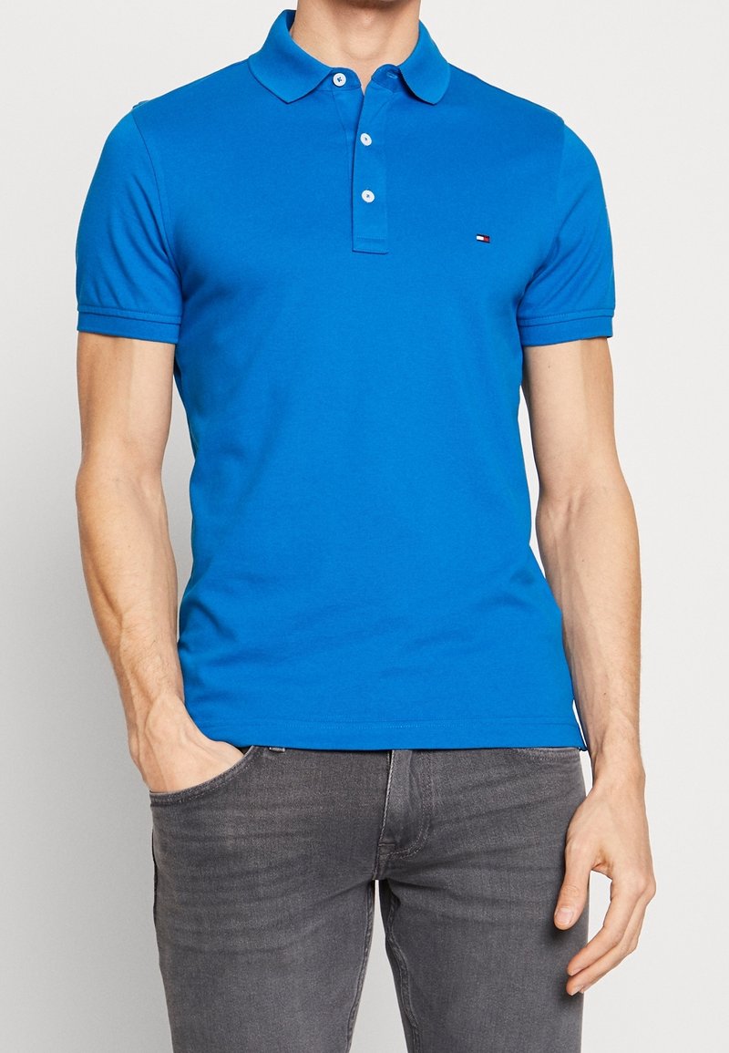 Chemise polo bleue en coton, dotée d'un col classique, de manches courtes et d'un petit logo sur la poitrine. Portée avec un jean gris.