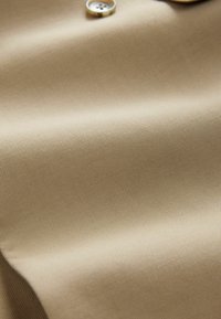 Tessuto beige con una texture liscia e un sottile motivo diagonale, caratterizzato da un bottone dorato. Ideale per applicazioni di abbigliamento.