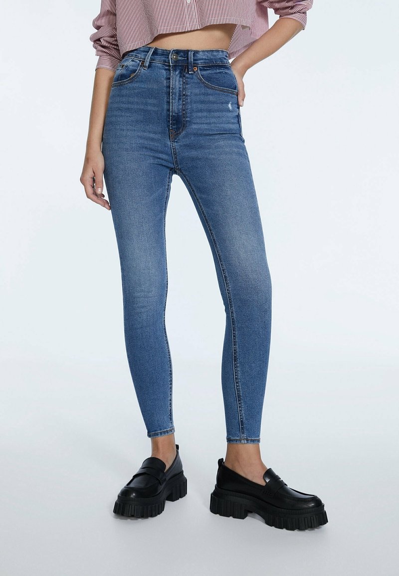 Femme portant un jean skinny bleu taille haute et des mocassins noirs épais à plateforme, avec une main posée sur la hanche.