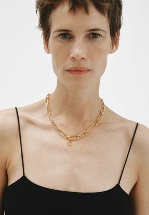 Mujer con cabello corto y castaño, que lleva un collar de eslabones dorados y una blusa negra de tirantes finos, sobre un fondo blanco.