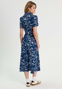 Blumenkleid aus navyfarbigem Stoff, kurze Ärmel, tailliert, fließender Rock. Hat hellblaue botanische Muster und weiße Sneaker mit schwarzen Akzenten.