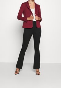 Een bordeauxrode strakke blazer met een enkele knoopsluiting, gedragen over een witte top en gecombineerd met zwarte uitlopende broek en zwarte sandalen met bandjes.
