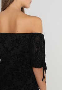 Vestido negro off-the-shoulder con textura de terciopelo floral, mangas de globo transparentes con lazos y una cintura ajustada.