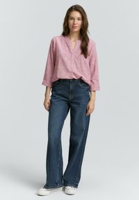 Chemise rose à manches longues avec un motif texturé, associée à un jean en denim bleu foncé à jambes larges et des baskets blanches. Tenue simple et décontractée.