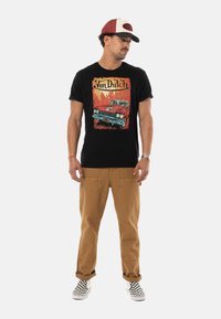 T-shirt noire avec un graphique de voiture vintage éclatant ; pantalon marron ; baskets à carreaux ; casquette de baseball avec des accents rouges.