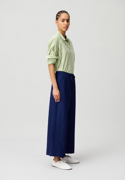 Camicia a righe verdi con maniche arrotolate, abbinata a pantaloni larghi blu navy e scarpe bianche. L'outfit presenta una vestibilità comoda e una consistenza liscia.