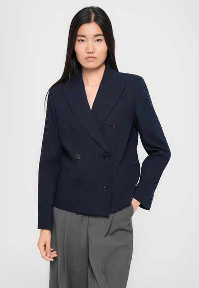 Blazer doppiopetto blu navy in un design strutturato, dotato di bottoni neri e rever a punte. Abbinato a pantaloni grigi plissettati.