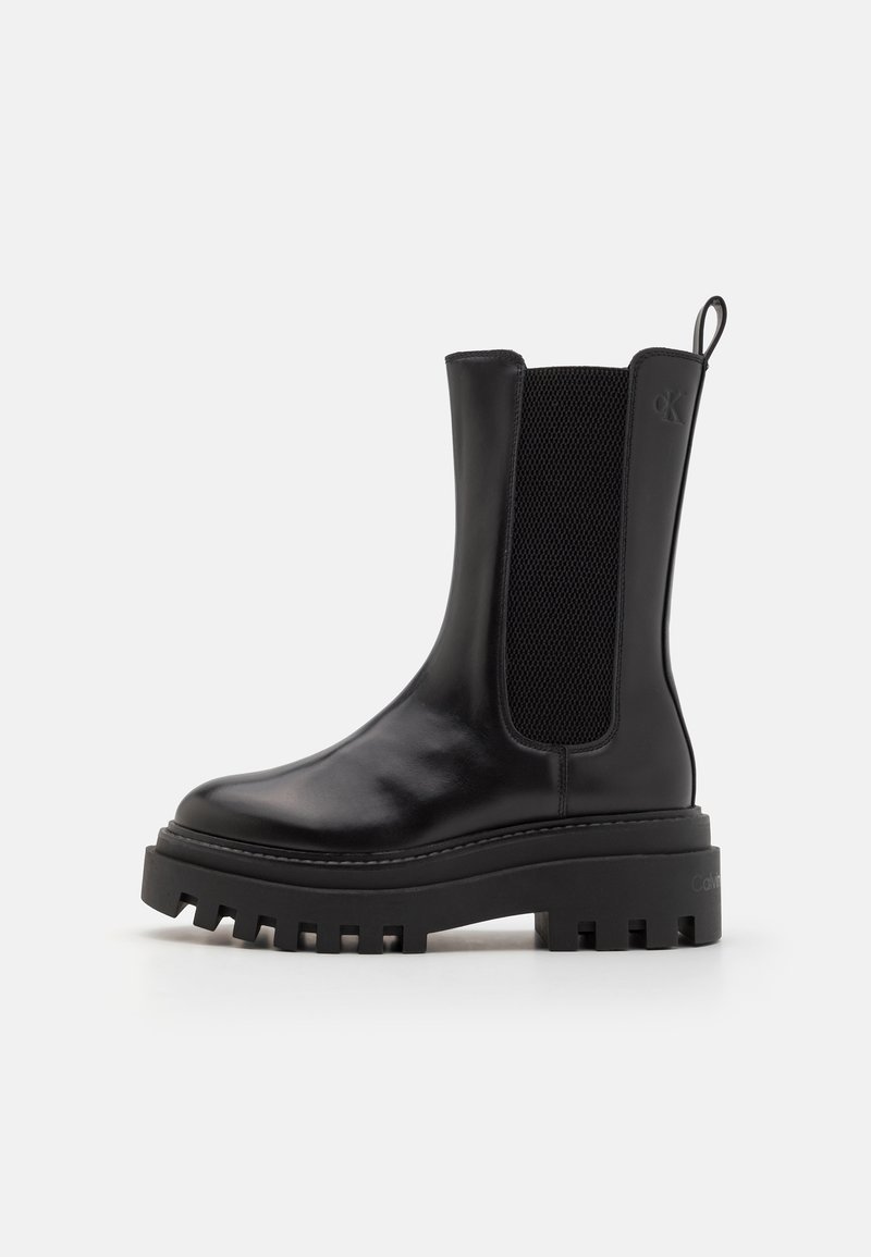 Bottes noires en cuir mi-mollet avec semelle crantée épaisse, panneaux latéraux élastiques et languette à l'arrière, arborant un logo discret sur le côté supérieur.