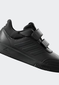 Chaussure de sport noire avec tige en cuir synthétique, accents texturés, deux bandes Velcro et trois rayures parallèles sur le côté.