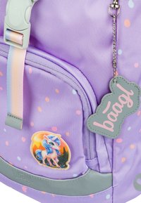 Zaino viola con motivo a pois, dettagli grigi, cerniera e un adesivo di unicorno. Include un'etichetta grigia con la scritta "baagl". Tessuto testurizzato.