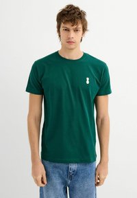 Camiseta de algodón verde con cuello redondo y mangas cortas. Presenta un pequeño logotipo de piña blanca en el lado izquierdo del pecho. Corte casual.