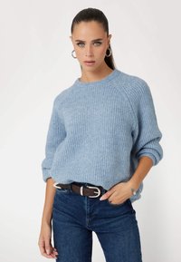 Maglione azzurro chiaro lavorato a maglia con scollo rotondo e maniche arrotolate, abbinato a jeans blu scuro e una cintura marrone con fibbia argentata.