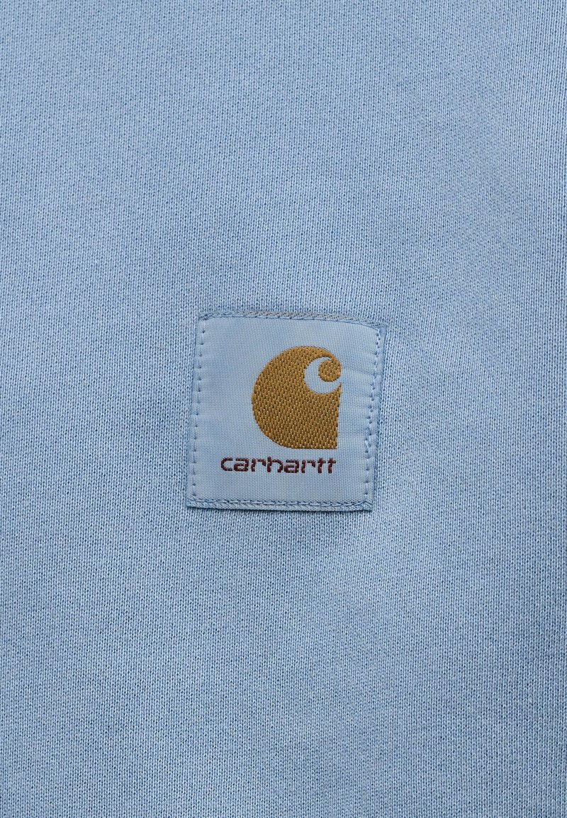 Hellblauer Stoff mit einem aufgenähten quadratischen Patch, der ein braunes, stilisiertes "C"-Logo und darunter das Wort "Carhartt" zeigt.