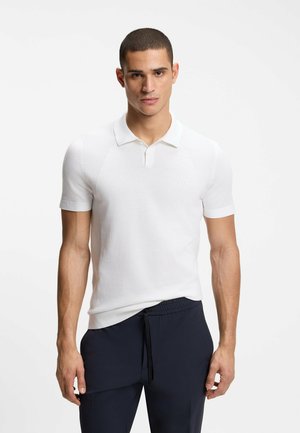 Mann mit kurzen Haaren trägt ein eng anliegendes weißes Kurzarm-Poloshirt und dunkle Jogginghosen mit Kordelzug, steht vor einem schlichten, hellen Hintergrund.
