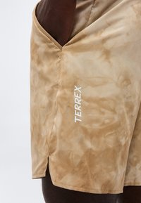 Μπεζ σορτς σε στυλ tie-dye με πλαϊνή τσέπη και λευκό λογότυπο "TERREX", φορεμένα από άτομο με το χέρι στην τσέπη.