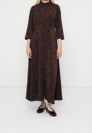 Frau trägt ein langes schwarzes Kleid mit dunkelroten Blumenmustern und dreiviertel Ärmeln, kombiniert mit glänzenden dunkelroten Ballerinas.