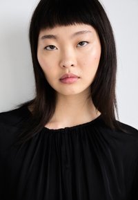 Musta, virtaava paita kerätyllä kaula-aukolla; sileä rakenne ja kevyt kangas; mallilla on suorat, tummat hiukset ja neutraali meikki.