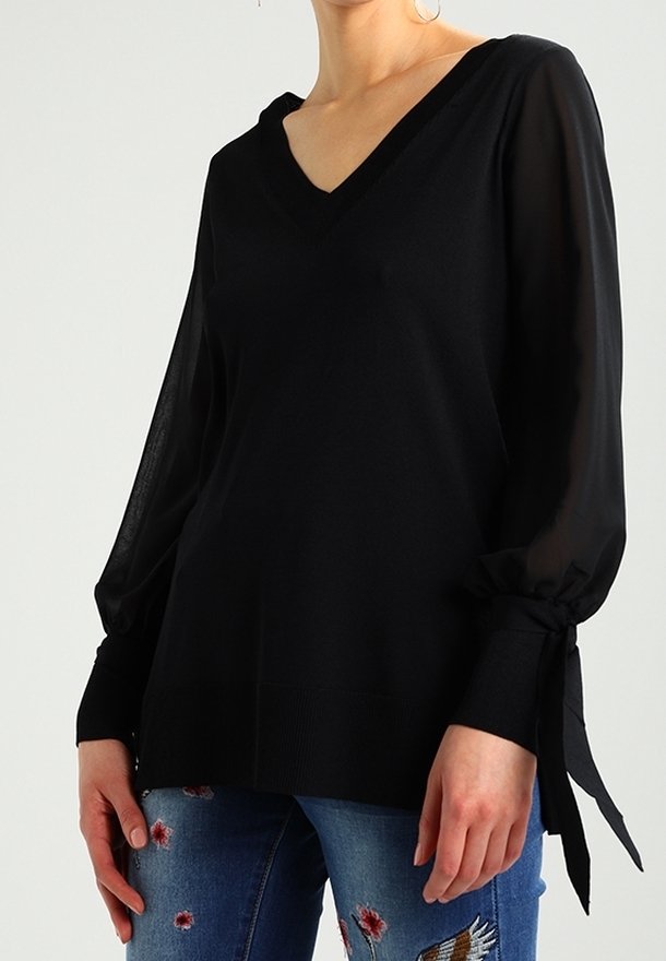 Czarny sweter z dekoltem w serek, długimi przezroczystymi rękawami i akcentami w postaci wiązań. Posiada dopasowane body i ściągacz na dole, noszony z haftowanymi niebieskimi dżinsami.