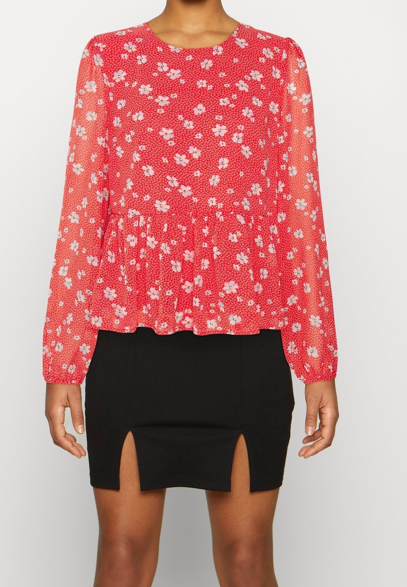 Blusa floreale rossa con maniche lunghe trasparenti, vita arricciata e mini gonna nera con due fessure laterali. Il materiale sembra leggero e testurizzato.