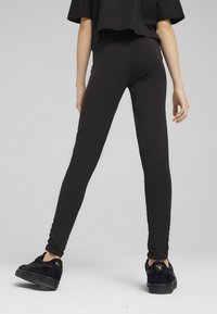 Černé legíny přiléhavého střihu s hladkým povrchem, v kombinaci s černými semišovými teniskami s zlatým logem, viděno zezadu.